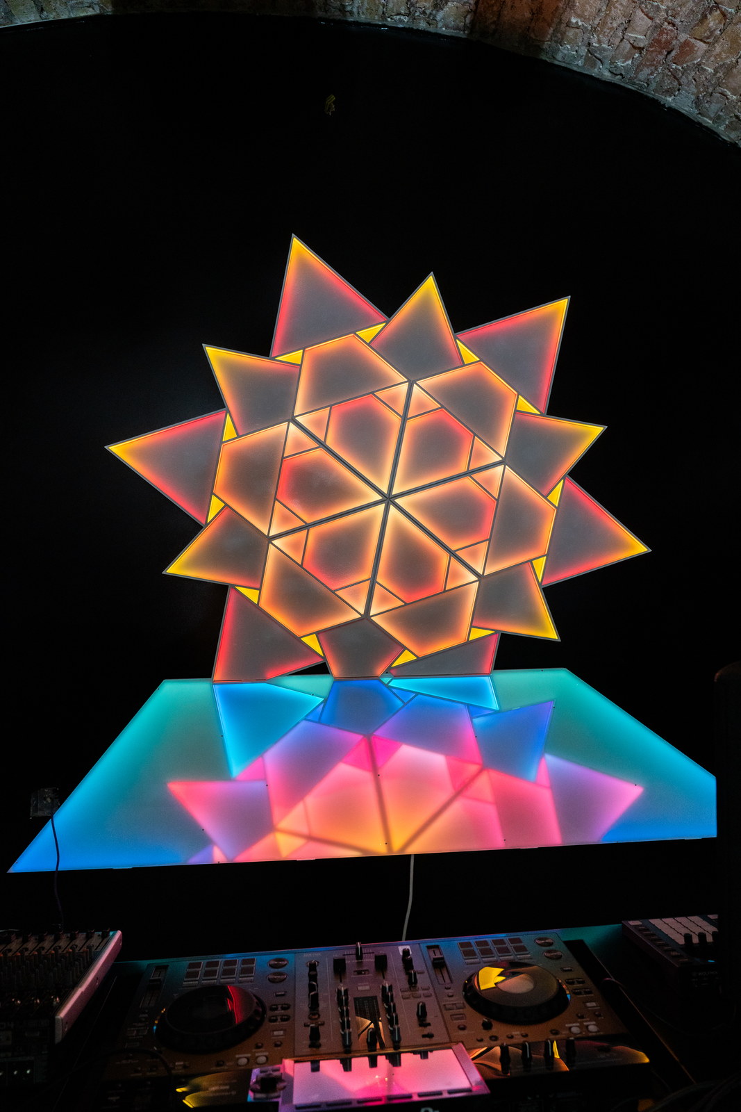 A star of backlit triangular panels glowing orange and pink on a black wall beneath a brick vault, above a lit counter of blue and pink glass.