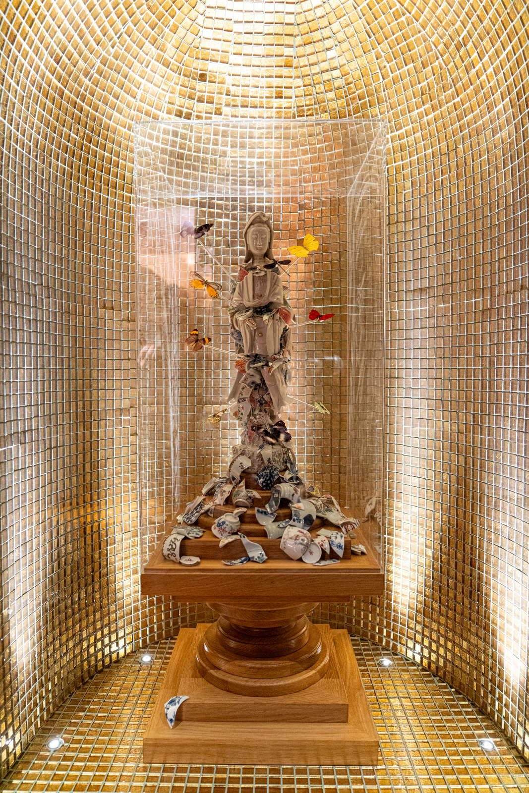The porcelain Guan Yin in her clear case seen closer, the gold mosaic of the niche curving behind her and the stepped wooden plinth below.
