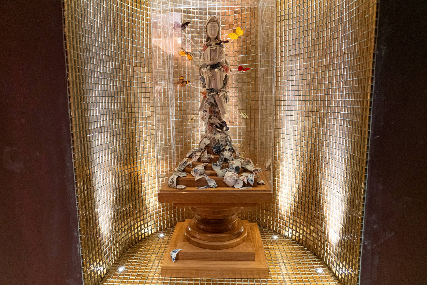 A porcelain figure of Guan Yin in a clear case on a turned wooden plinth, butterflies on thin stems around her, in a niche of gold mosaic lit from below.