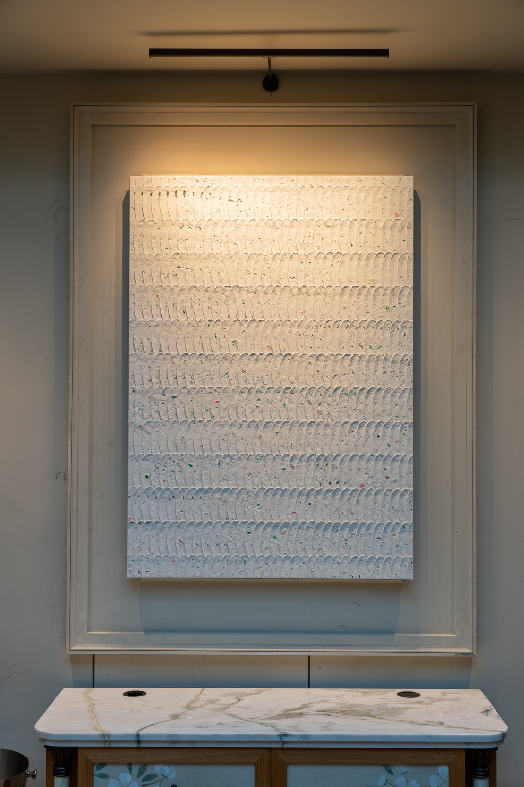 A white canvas worked in close rows of ribbed impasto, lit from above in a pale panelled frame on the wall, a marble-topped console below.