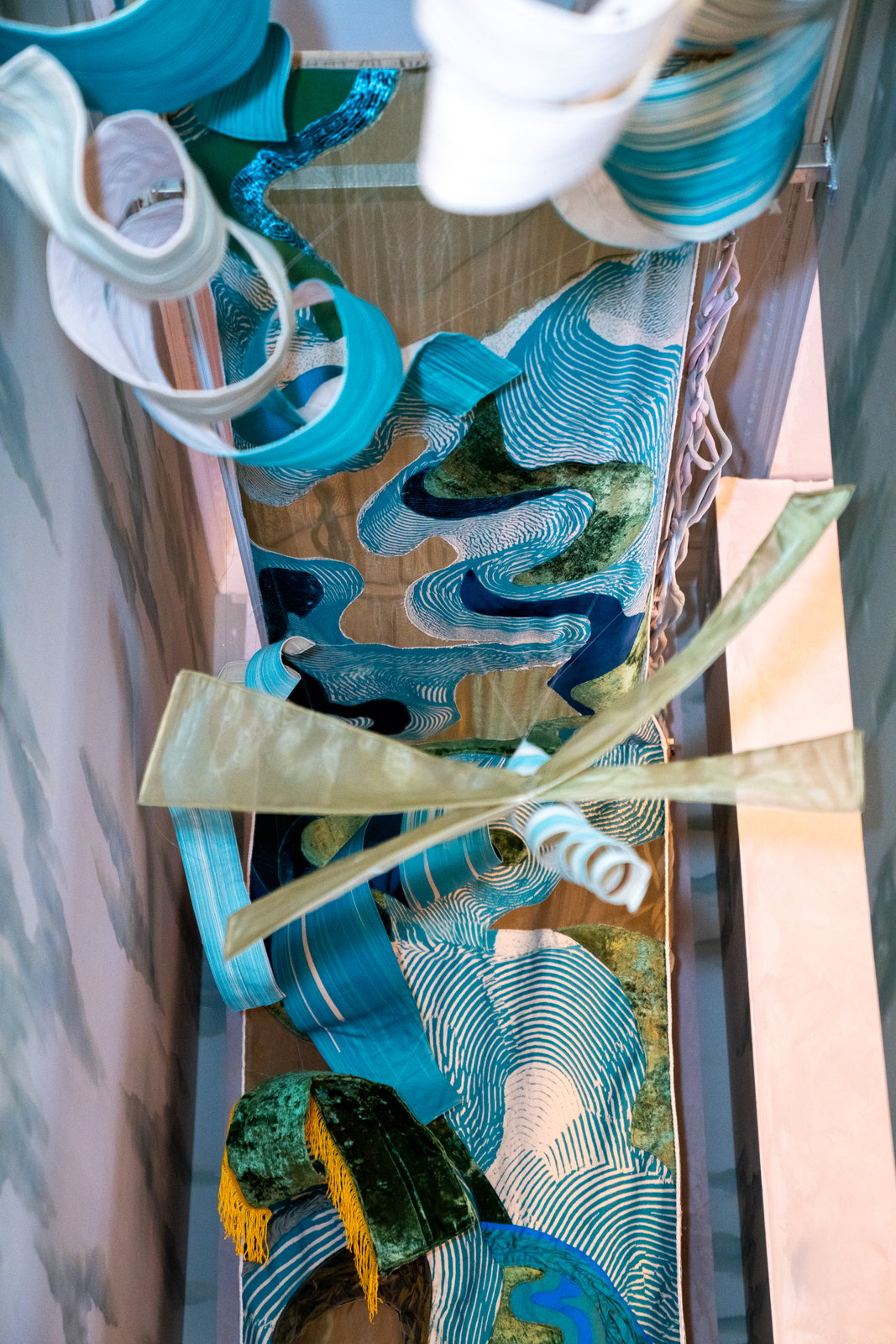 Lengths of blue, white and green printed fabric and twisted ribbon suspended in a narrow stairwell, seen from below.