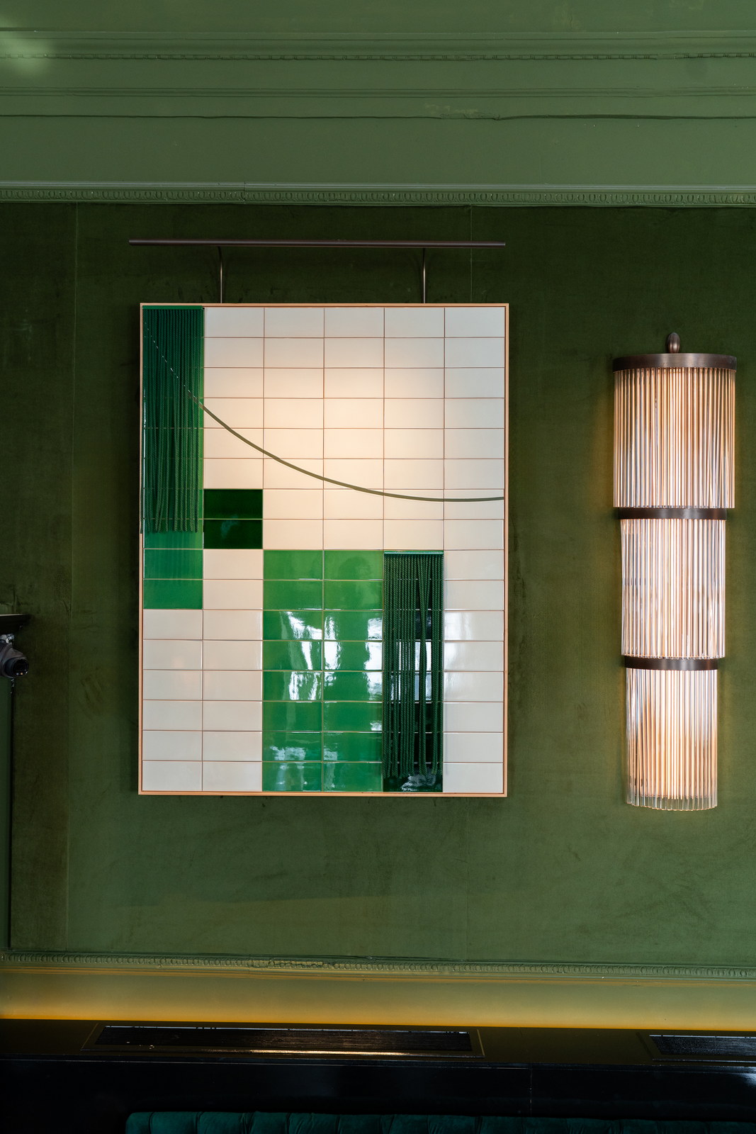 The tiled panel of cream and emerald green hung from its bronze rod on the green velvet wall, a ribbed wall light at its right.