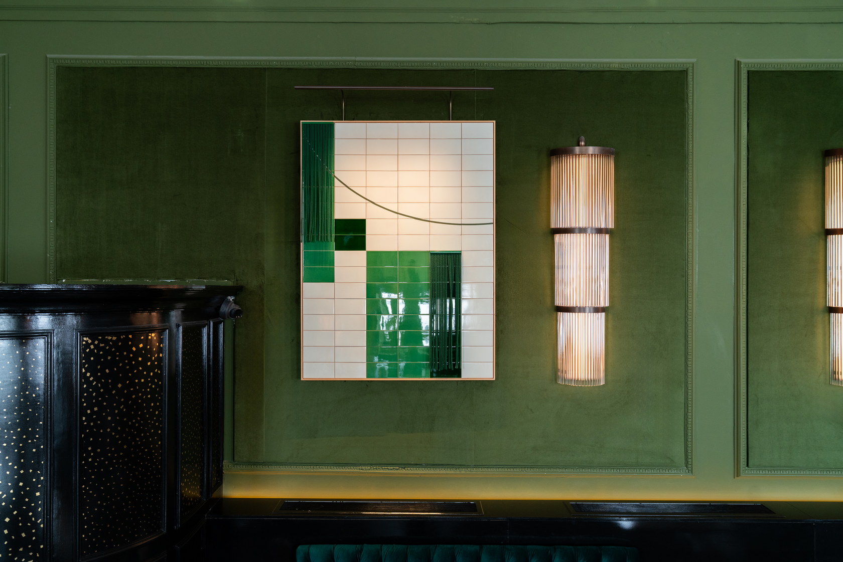 A framed panel of cream and emerald-green glazed tiles with a dark green fringe, hung from a bronze rod on a green velvet wall beside a ribbed wall light.