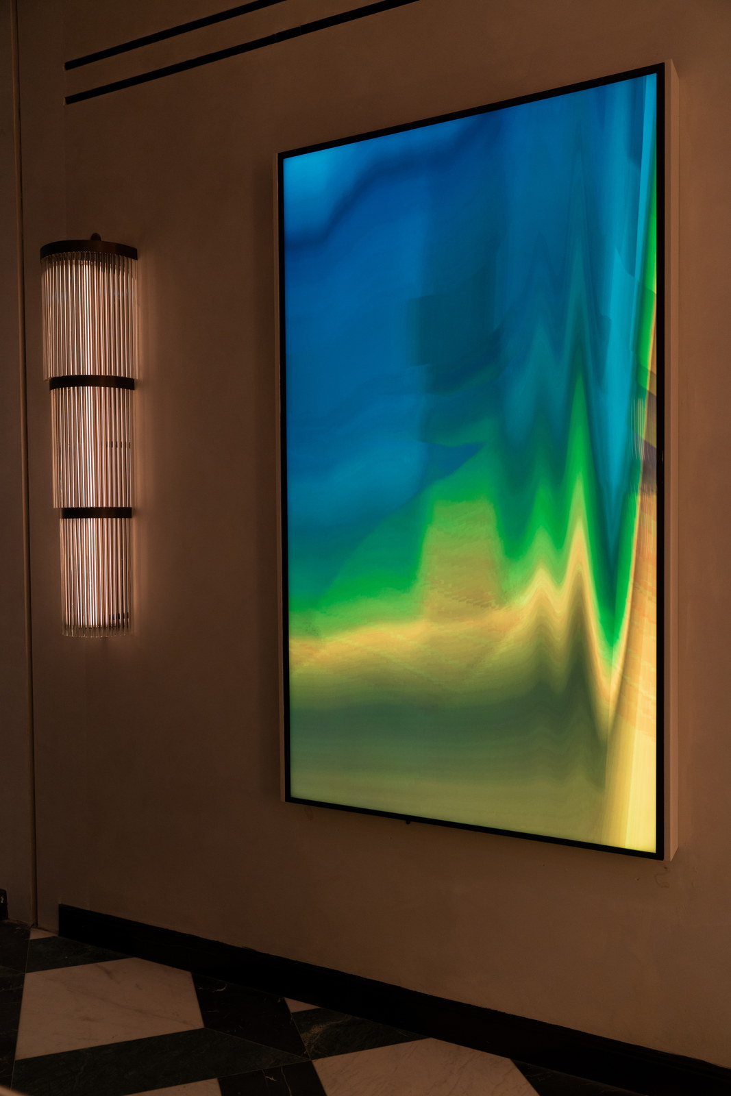 A tall framed screen glowing blue, green and gold on a pale wall at the end of a corridor, a ribbed wall light at its left.