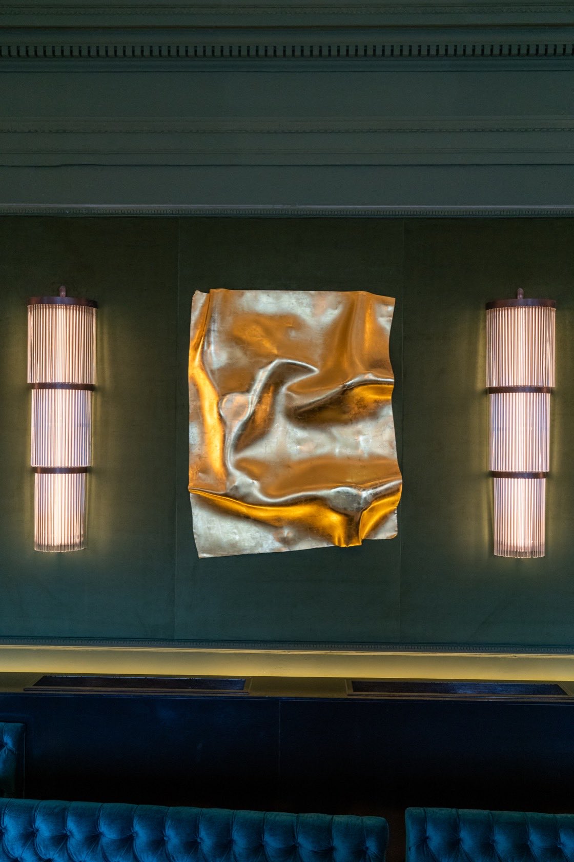 The same folded metal sheet seen from lower down, the panelled cornice above it and a buttoned banquette running beneath.