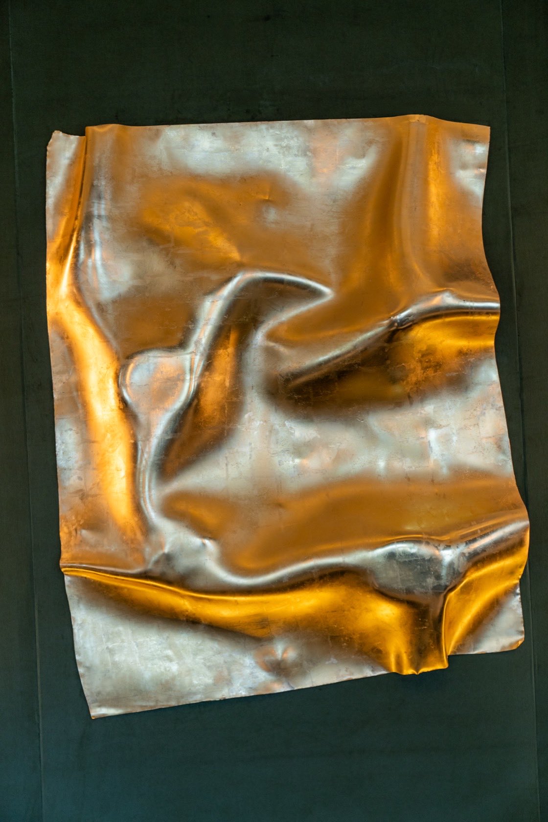 The folded metal sheet close up, its surface creased into soft ridges that hold gold at the folds and silver in the flats.