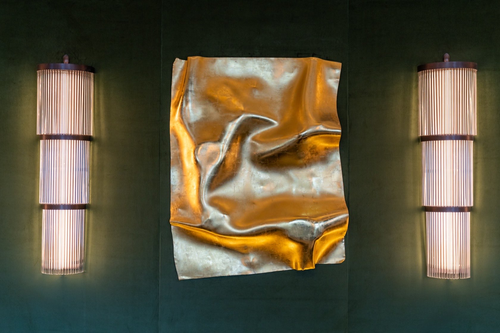A folded sheet of metal mounted on a dark green wall, its creases catching gold and silver light, flanked by two tall ribbed wall lights.