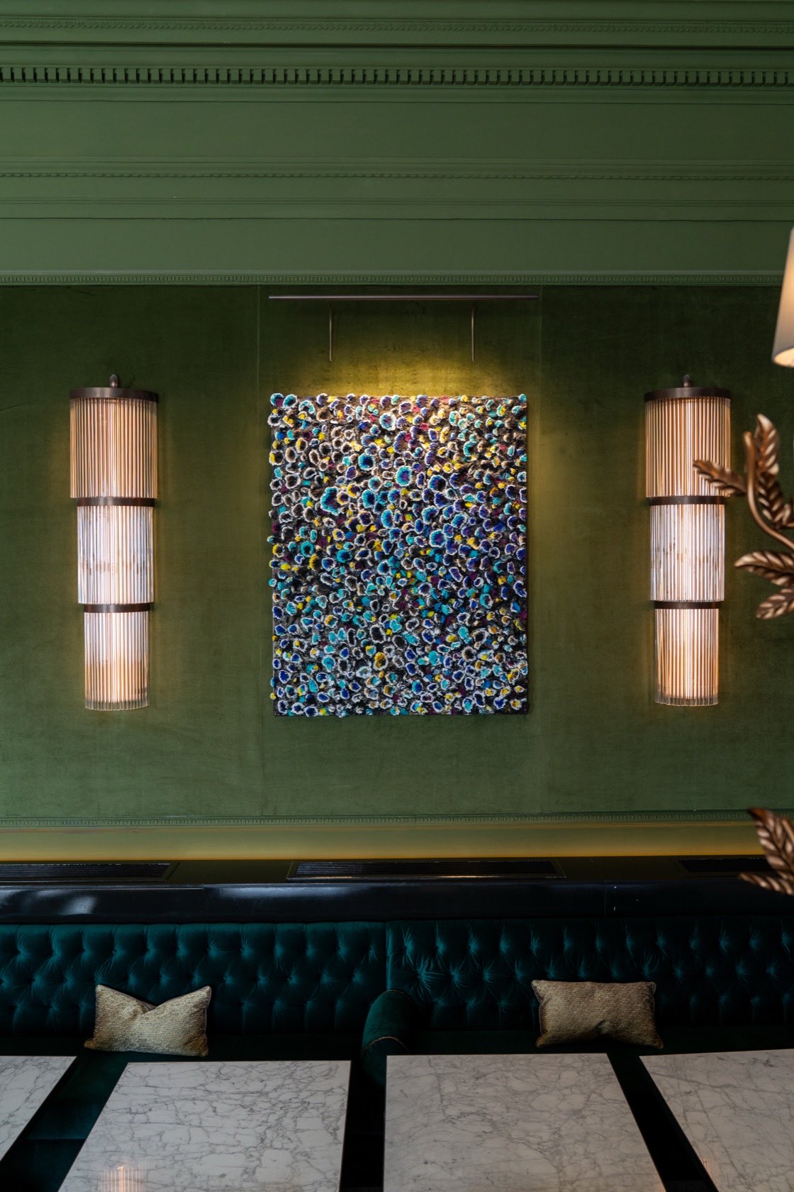 A panel of dense blue, yellow and white rosettes on a green velvet wall between two ribbed wall lights, above a buttoned banquette and a marble floor.