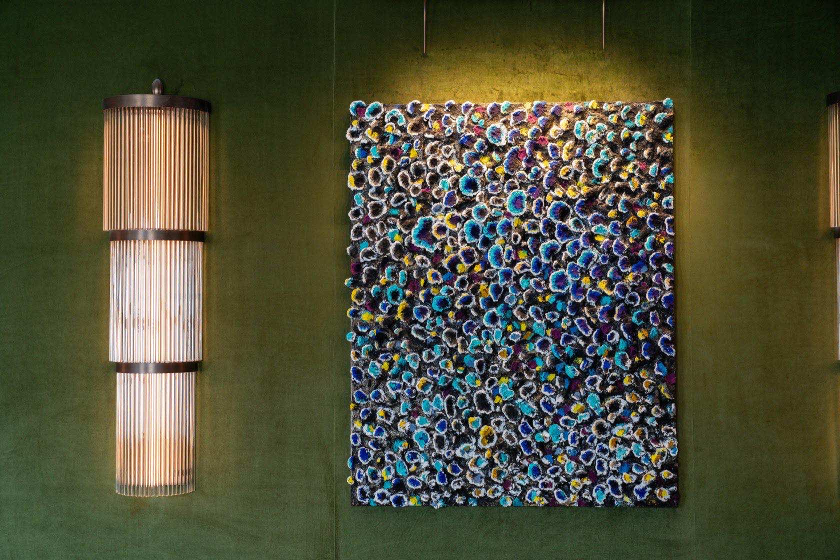 A panel of dense blue, yellow and white rosettes on a green velvet wall, one ribbed wall light at its left, seen at the scale of the room.