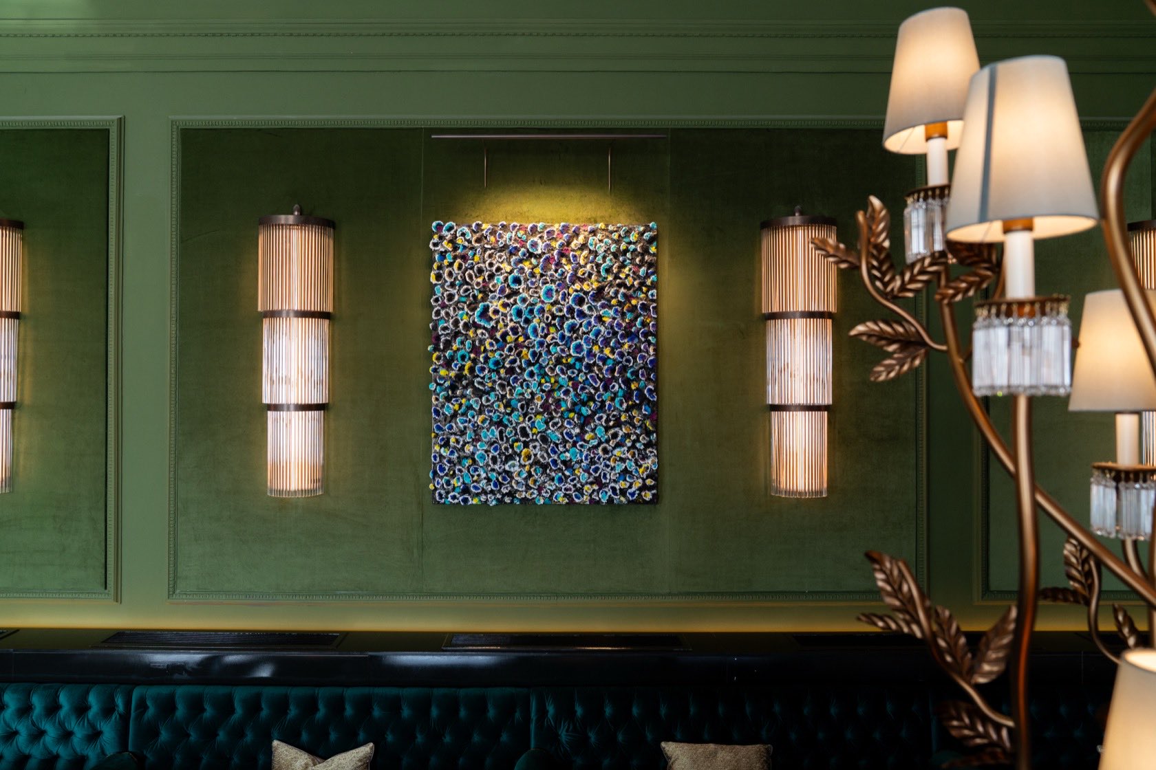 A panelled green dining room: a densely textured panel lit from above between two ribbed wall lights, a buttoned banquette below and a branched candelabrum at the right edge.