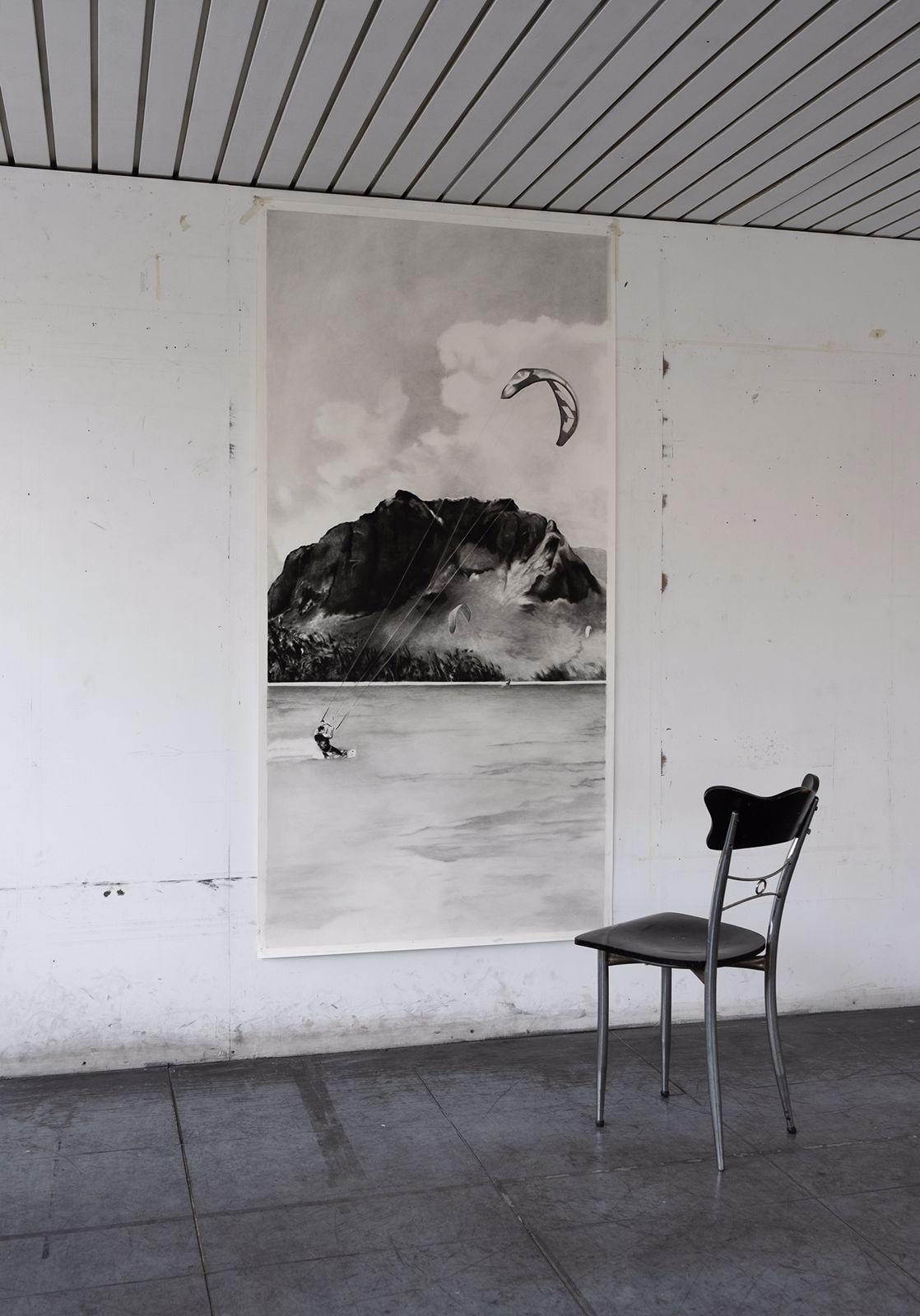 A tall grey drawing of a kite surfer on flat water below a dark headland, taped to a white wall beside one metal chair.