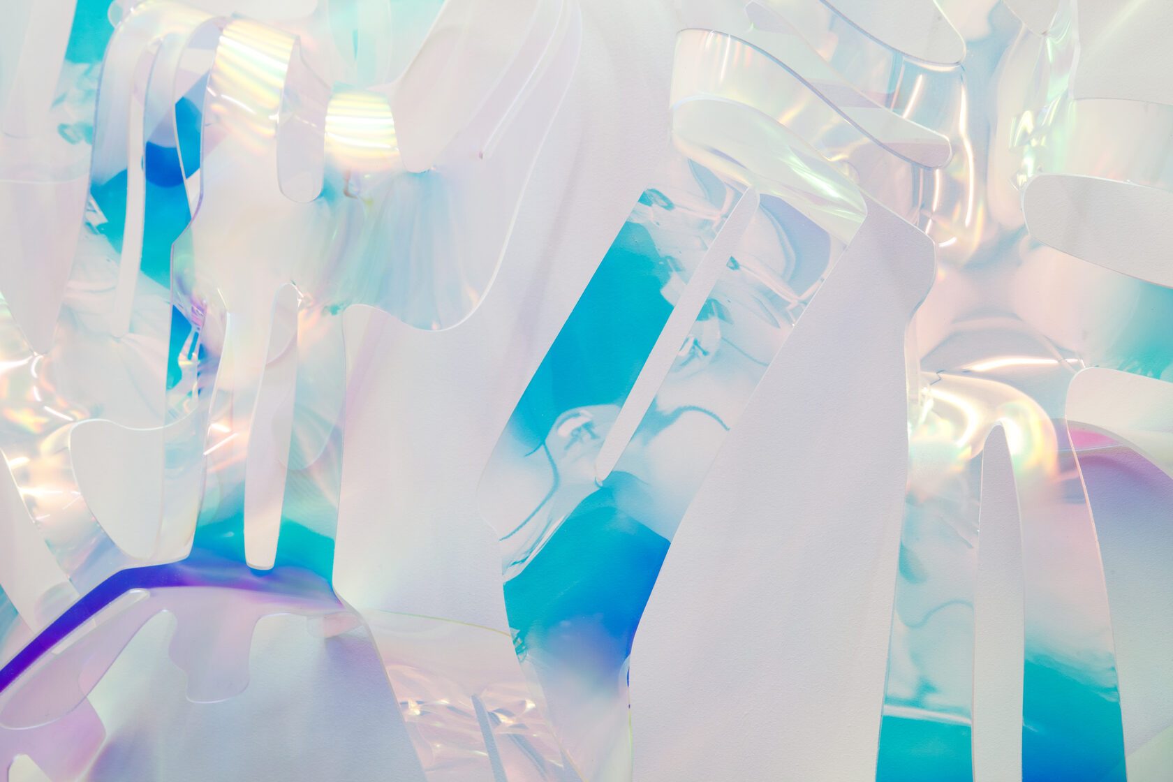 A detail of a sculpture: overlapping translucent strips of acrylic glass in blue, green and clear, filling the frame against a white wall.