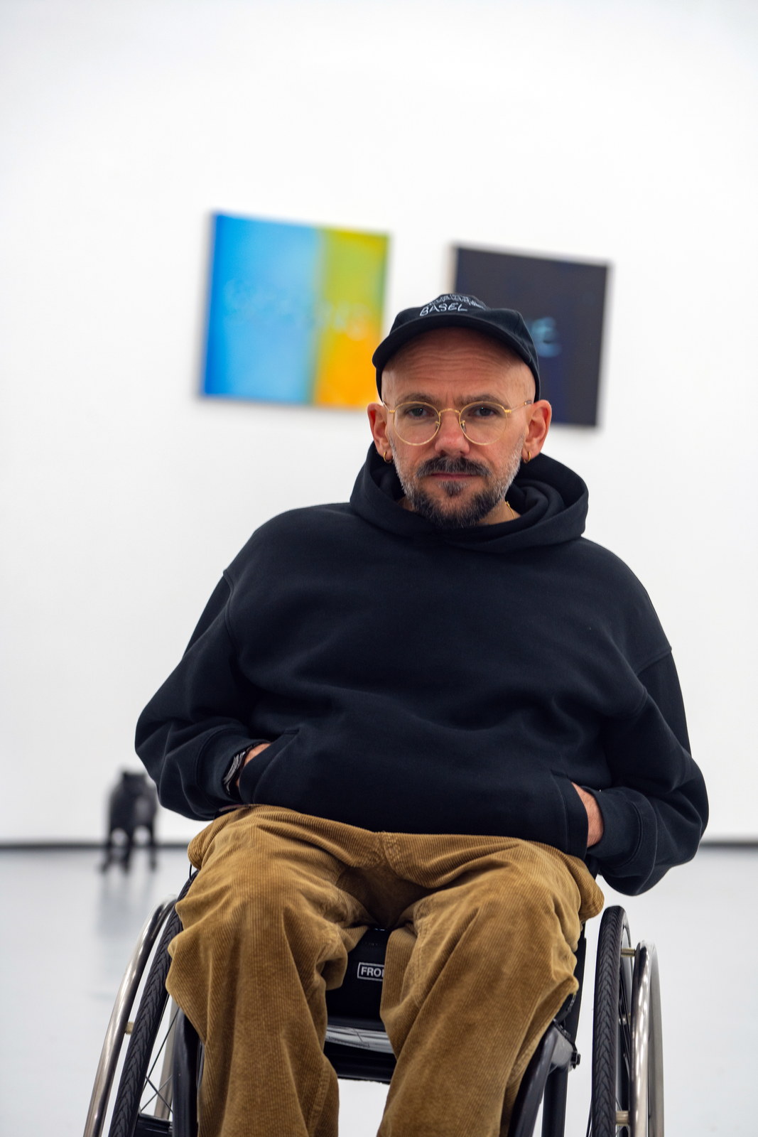 The artist in the gallery with two bright panel works on the wall behind him and a black cat standing at his side.