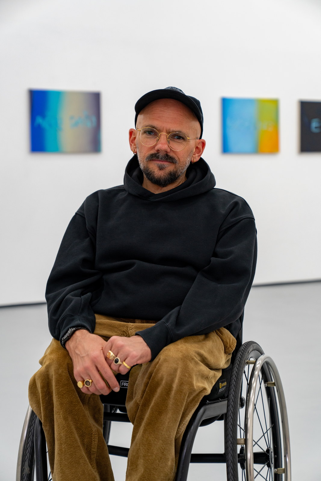 The artist seated in a wheelchair in a white gallery, four small bright panel works spaced along the wall behind him.