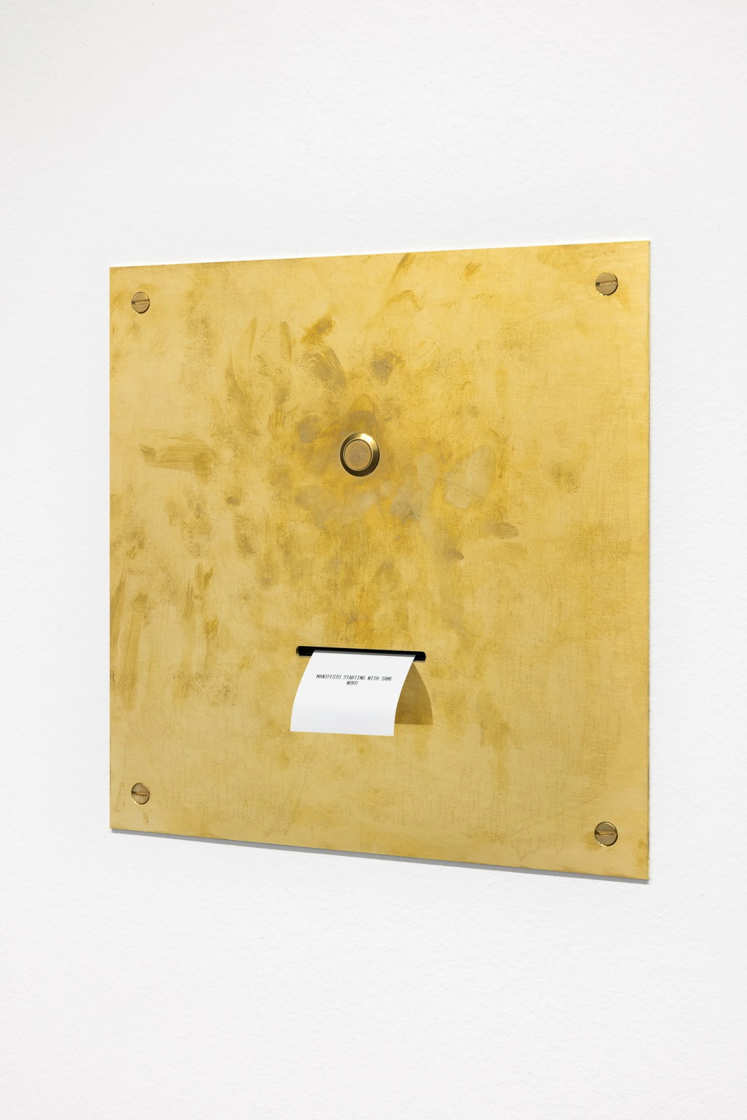 A square brass panel fixed flush to a white wall with four screws, a round button at its centre, and a slot below it from which a white printed slip curls out.