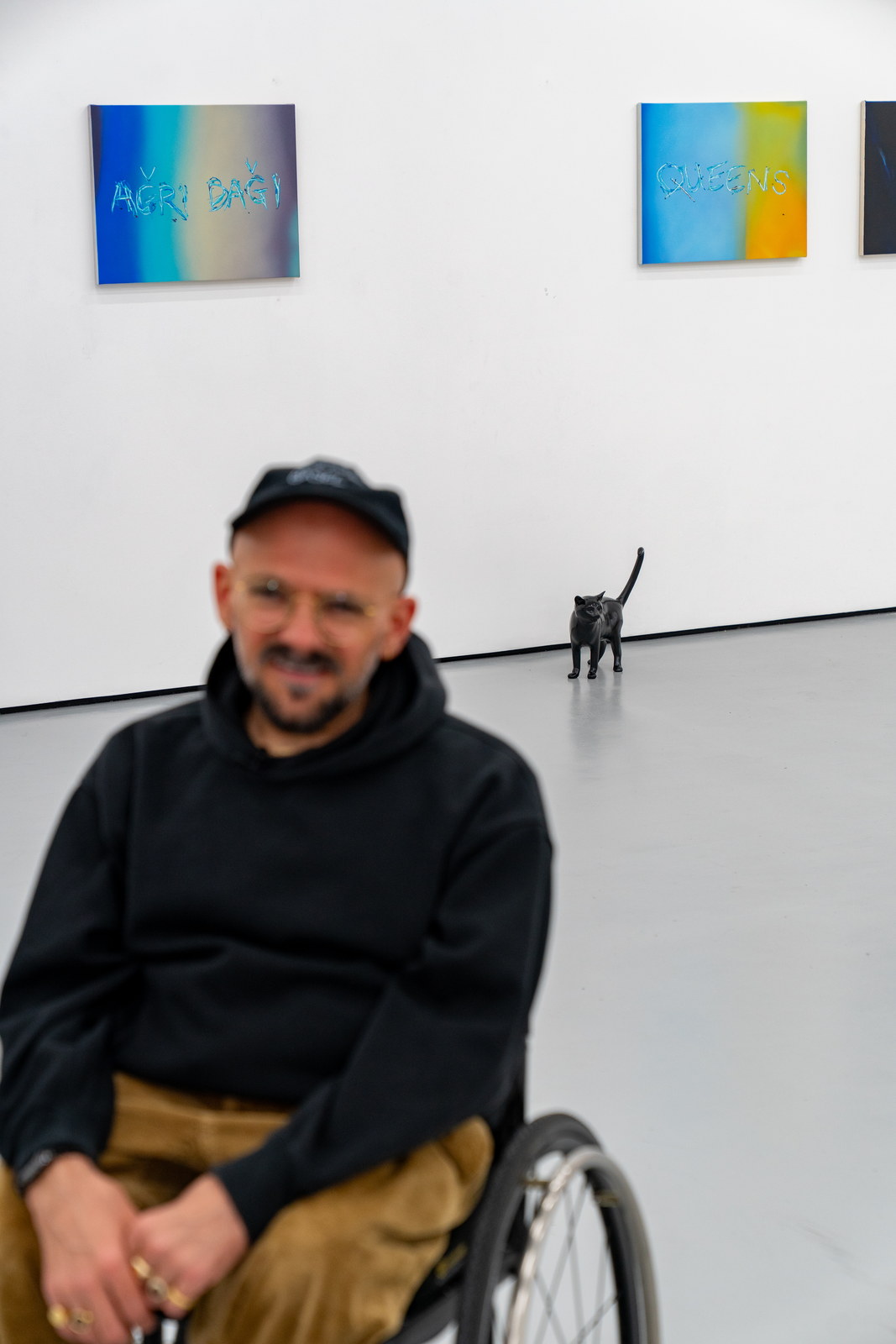 The artist seated in the foreground, out of focus, the same gallery behind him: panel works along the wall and a black cat on the floor.
