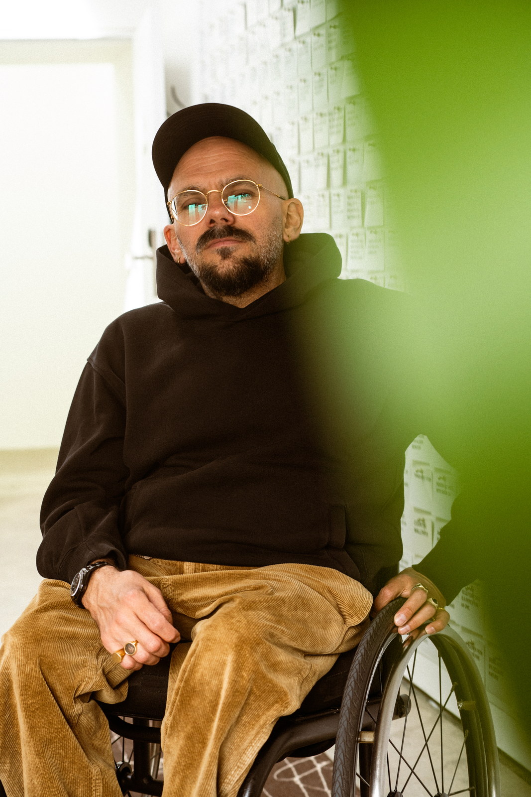 The artist in his wheelchair beside a pale wall, green light falling across it from the right.
