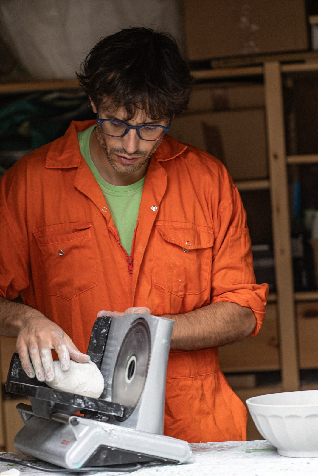 The artist at work in his studio, an electric slicer in front of him holding a block of white clay, a white bowl at his side.