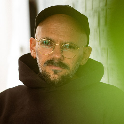 Ryan Gander, portrait: a man in a dark cap and hooded sweatshirt with round metal-framed glasses, turned to the camera, green light falling across the wall behind him.
