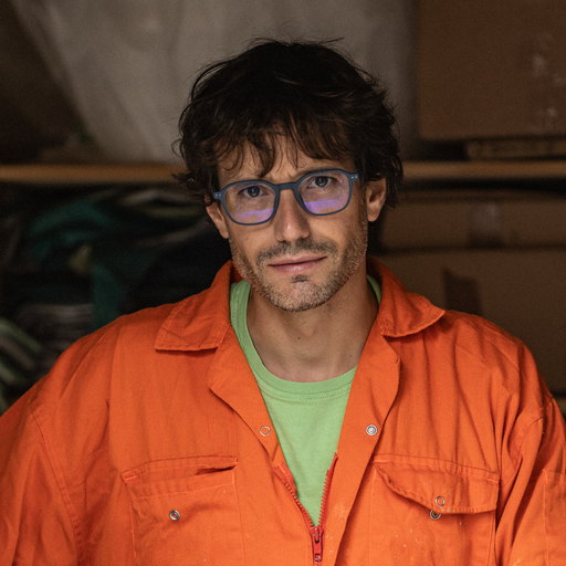 Albano Hernández, portrait: a man in an orange work jacket over a green shirt, with dark curled hair and blue-framed glasses, standing in his studio.